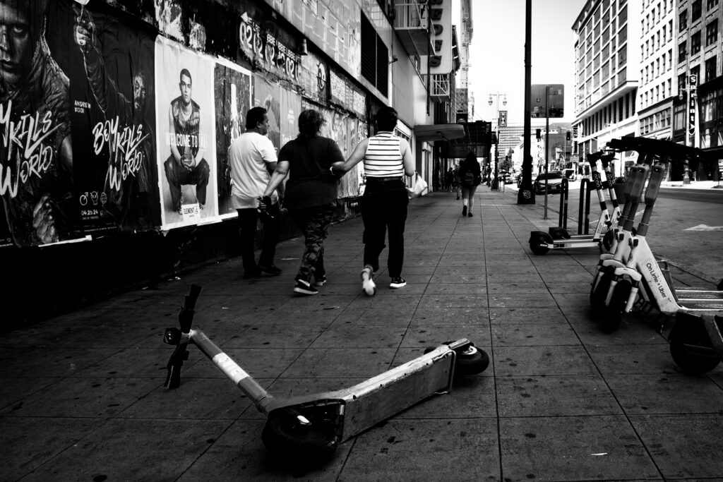 A black and white photo of a sidewalk in downtown Los Angeles. A Bird Scooter is laying across the sidewalk. in the background, three people are walking away. More scooters, a wall covered with posters, and office buildings are in the background.