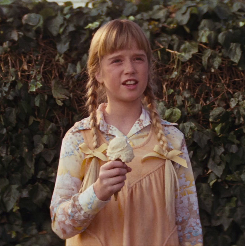 Kim Richards, roughly age 11, with pigtails that end with yellow bows, stands holding an ice cream cone in her right hand. The background is ivy.