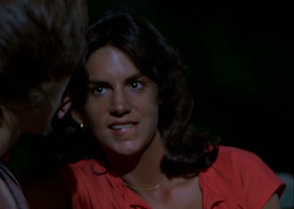 Mercedes Ruehl  as an undercover policewoman at night, wearing a red shirt, biting her lip and looking at a partially visible Ajax in The Warriors.