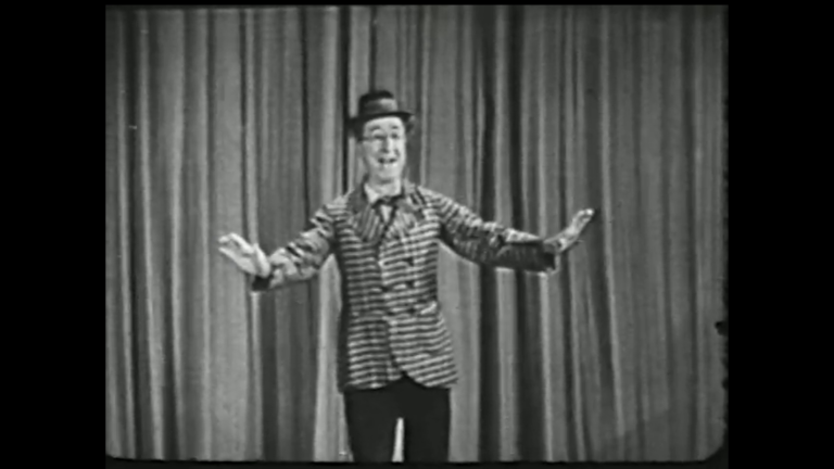 Black and white still from a tv show. Ed Wynn, wearing a black and white striped jacket and hat, standing in front of a stage curtain.