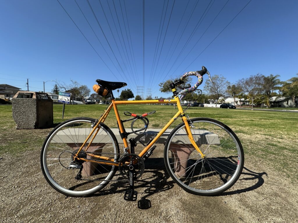 My bike. A Surly Long Haul Trucker. The frame fades color from yellow to orange. The handlebars are wrapped with multicolor bar tape. the frame has a U-lock and a cable attached to it.

The bike is in a park on a clear day, leaning against a cement bench. A trash can and a sign are visible in the background. The bike is sitting on a dirt circle with very short yellow and green grass around it. There are trees and cars in the distance. The sky is clear blue. Power lines overhead recede into the distance.