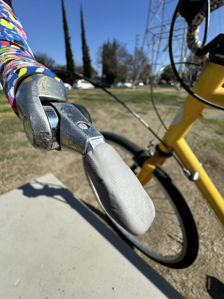 A close-up of a bar-end shifter. The shifter is scraped and worn, and has a lumpy gray blob covering the shift handle.

The handlebar (wrapped with multicolor bar tab) and the front of a yellow bicycle are visible. Out of focus in the background are a park with an electrical tower, some trees, and some cars.