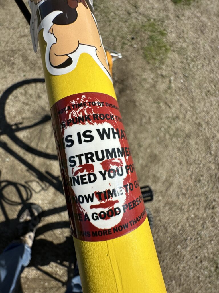 The top tube of a yellow bicycle, shot from above. The tube has two visible stickers: the bottom half of a drawing of Porky Pig, and a red stick with a white portrait of Joe Strummer superimposed with a partially visible quote from Henry Rollins - the edges wrap around and hdes the first and last last letters of each line.
The full quote is:
THIS IS NOT A TIME TO BE DISMAYED
THIS IS PUNK ROCK TIME
THIS IS WHAT
JOE STRUMMER
TRAINED YOU FOR
IT IS NOW TIME TO GO
YOU'RE A GOOD PERSON
THAT MEANS MORE NOW THAN EVER

What's actually visible is:
S NOT A TIME TO BE DISMA
S IS PUNK ROCK TIM
IS IS WHAT
E STRUMMER
INED YOU FOR
NOW TIME TO GO
RE A GOOD PERSON
EANS MORE NOW THAN EVER