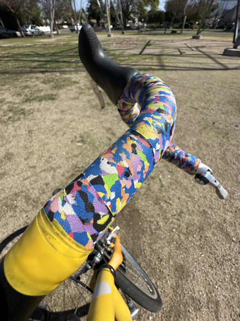 The handlebar (wrapped with multicolor bar tab) and the front of a yellow bicycle are visible. Out of focus in the background are a park, some trees, and some cars.