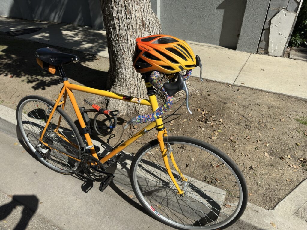 My bike. A Surly Long Haul Trucker. The frame fades color from yellow to orange. The handlebars are wrapped with multicolor bar tape. the frame has a U-lock and a cable attached to it. 

The bike is standing up in a cement gutter. A yellow, orange and black Specialized helmet rests on the handlebars.