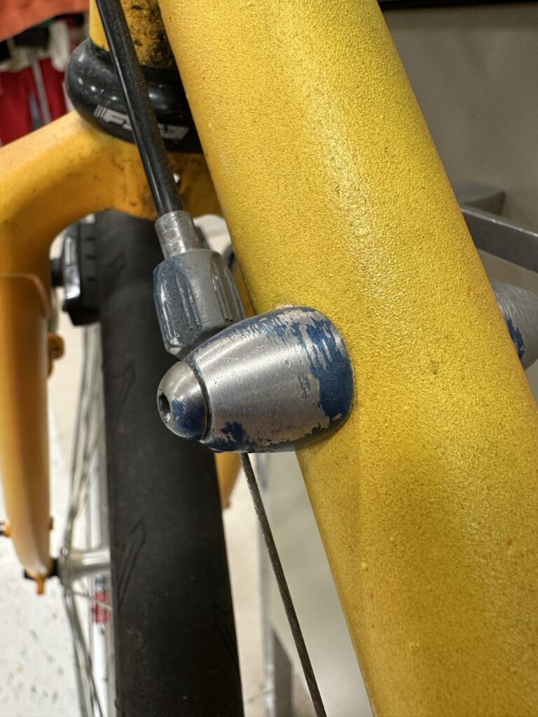 Closeup of a cable guide on the frame of my yellow bike. The guide is silver, with blue paint partially scraped off.